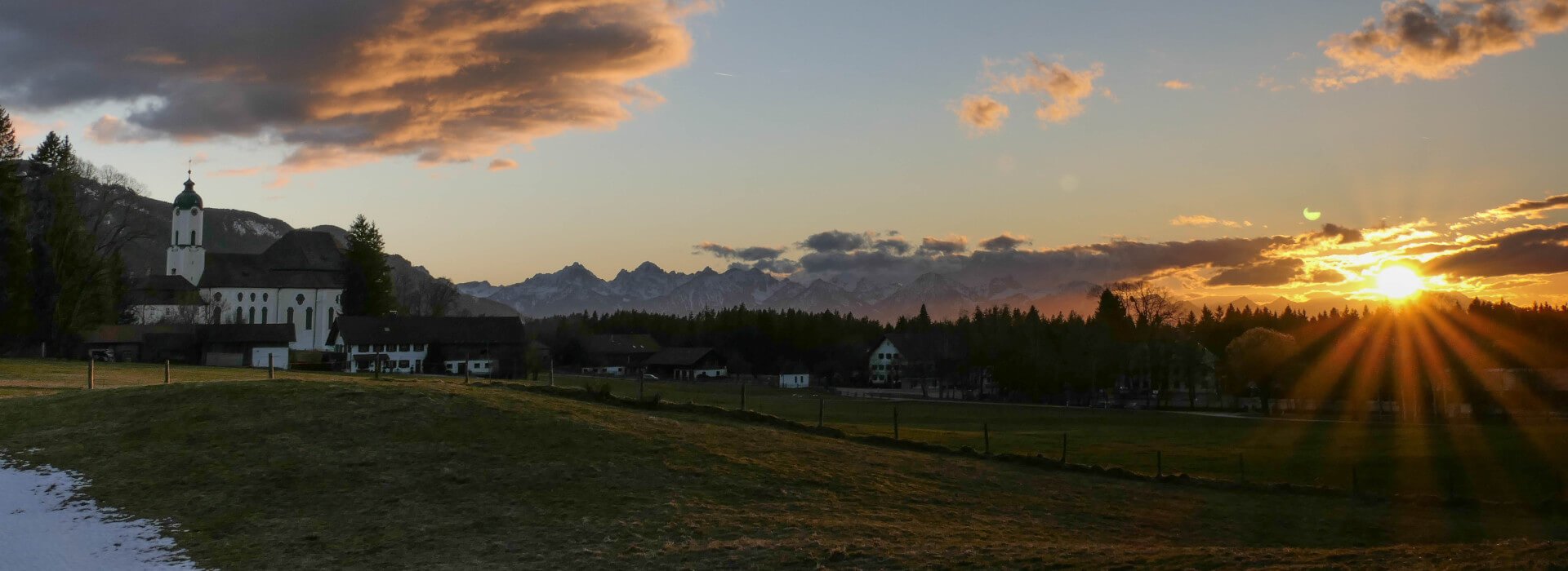 Sonnenuntergang über der Gemeinde Wildsteig. Die Sonne wirft lange Schatten und im Hintergrund erstrecken sich die Alpen.